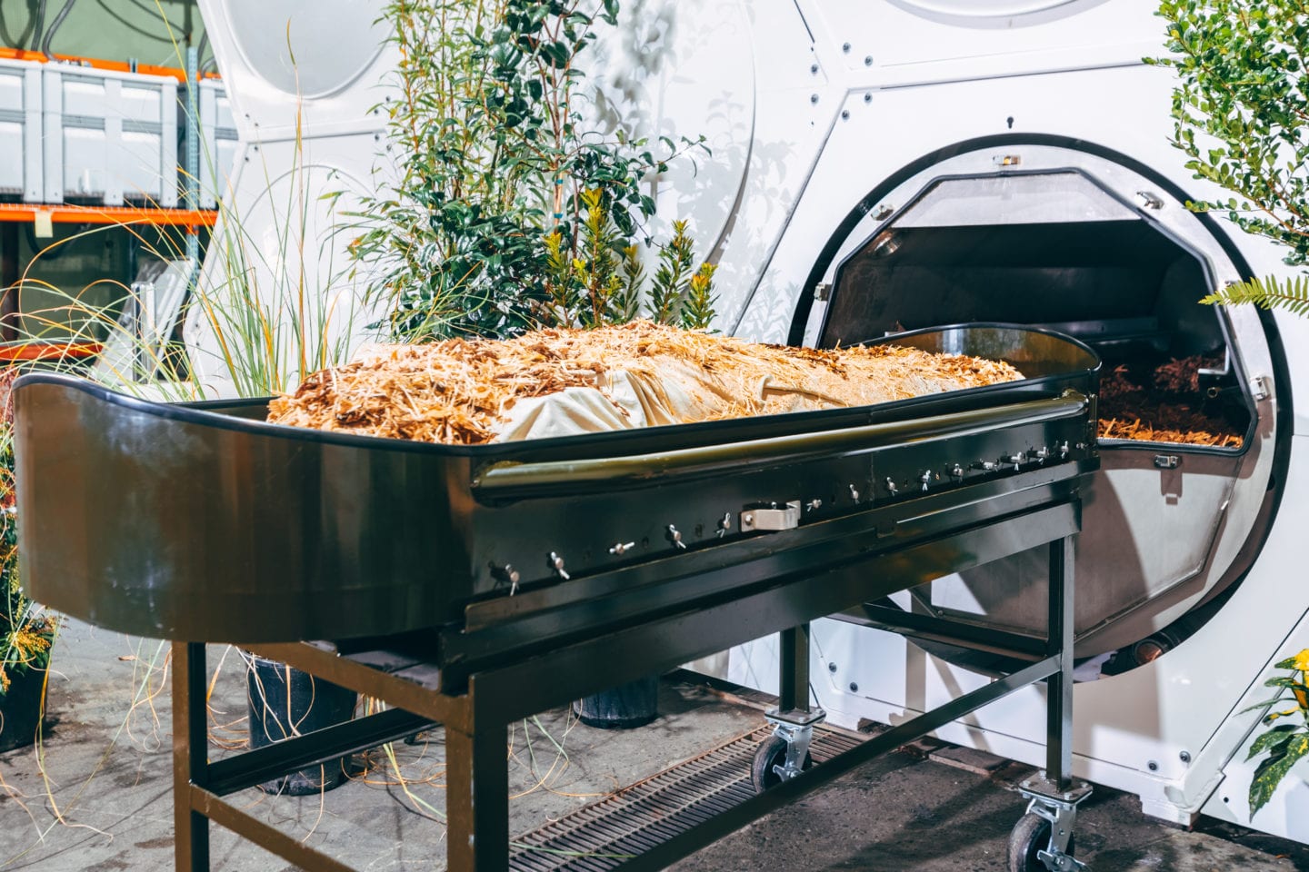 A mannequin covered in plant material rests on a cradle in front of an open vessel.